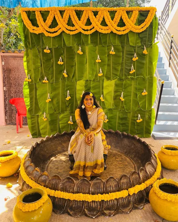 A unique Haldi setup featuring a wall of fresh banana leaves, marigold garlands, and a large brass Urli for the bride, creating a very traditional South Indian look.