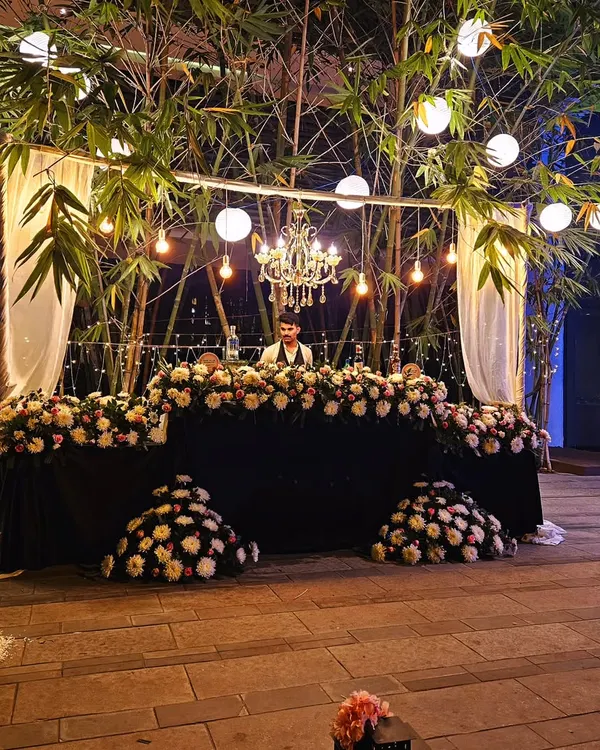 An elegant outdoor bar setup nestled among bamboo trees. The bar is heavily decorated with white and green florals, with a chandelier and string lights overhead for a magical touch.
