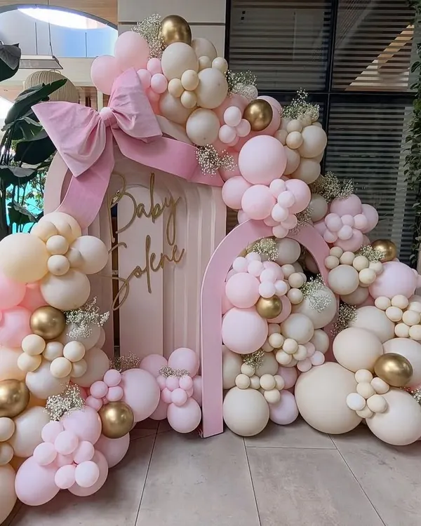 An elaborate and beautiful baby shower setup. The double arch backdrop is covered in an organic arrangement of pink, cream, and gold balloons, with a large pink bow as the finishing touch.