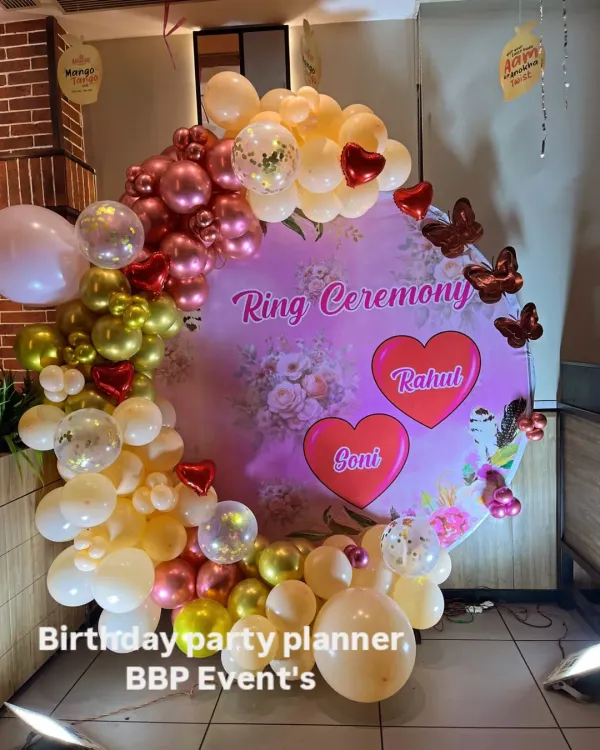A personalized ring ceremony backdrop with a custom printed floral design featuring the couple's names, 'Rahul' and 'Goni'. The ring is adorned with a balloon arch in shades of pink, peach, and gold.