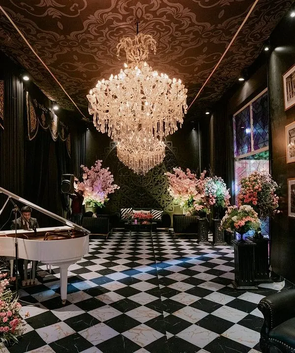 The entrance to a chic pre-wedding event, featuring a grand white piano on a black-and-white checkered floor. The room is filled with crystal chandeliers and soft pink floral arrangements, creating a stunning contrast with the dark, patterned ceiling.