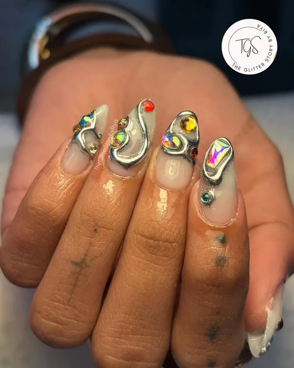 An edgy and artistic set featuring sculpted silver elements. Each nail has a unique abstract design, accented with colorful crystals for a pop of color against the metallic finish.