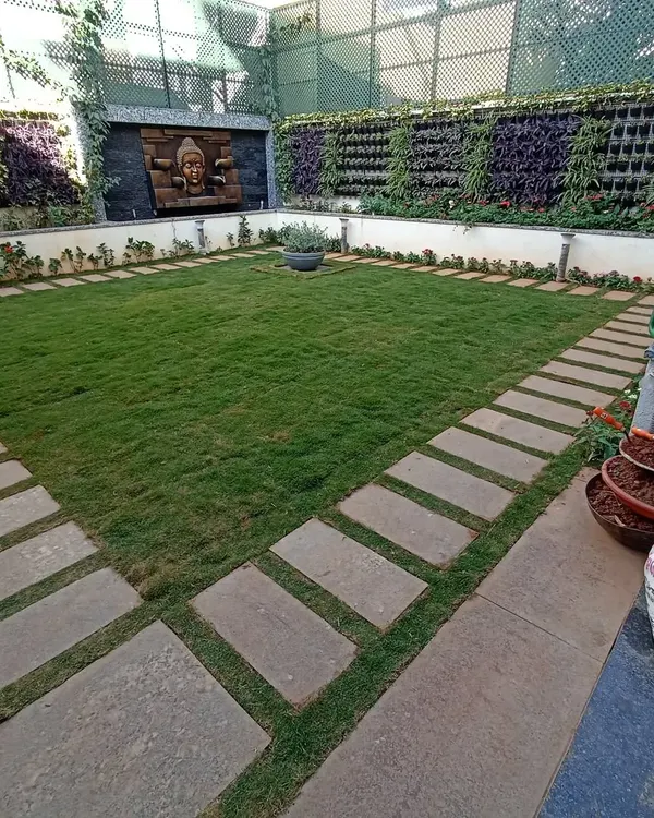 A formal courtyard garden featuring a central lawn bordered by stone pavers. The space is enclosed by lush vertical gardens and a serene Buddha water feature, creating a private and symmetrical oasis.