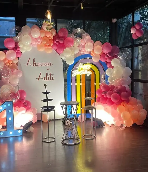 A beautiful pastel-themed first birthday party, featuring a large balloon arch, a rainbow-colored door prop, and a prominent light-up number '1'.