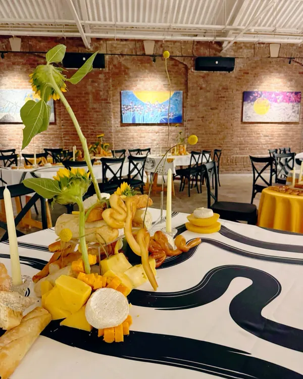 An art gallery event where the tablescape itself is a work of art. This centerpiece is crafted from an assortment of cheeses, breads, and sunflowers, set against an exposed brick wall and modern paintings.