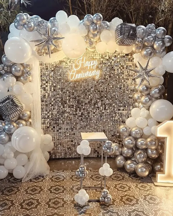We create stunning, photo-ready backdrops for any milestone. This first-anniversary celebration features a silver shimmer wall, a custom "Happy Anniversary" neon sign, and a large light-up number, all framed by a beautiful silver and white balloon arch.