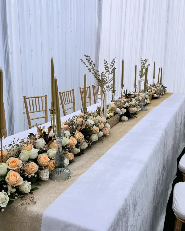 A full view of a long dining table dressed in an elegant floral arrangement. The design features a burlap runner topped with a dense mix of peach and white roses, accented with tall golden candles for a romantic and sophisticated dinner setting.