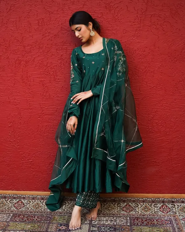 A moment of quiet grace. The deep emerald of the anarkali against the bold red backdrop creates a visually rich and sophisticated portrait.