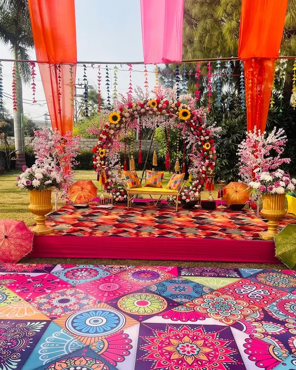 A vibrant Haldi and Mehendi stage setup with a beautiful floral swing. The colorful printed flooring and bright drapes create a festive atmosphere perfect for an outdoor celebration.