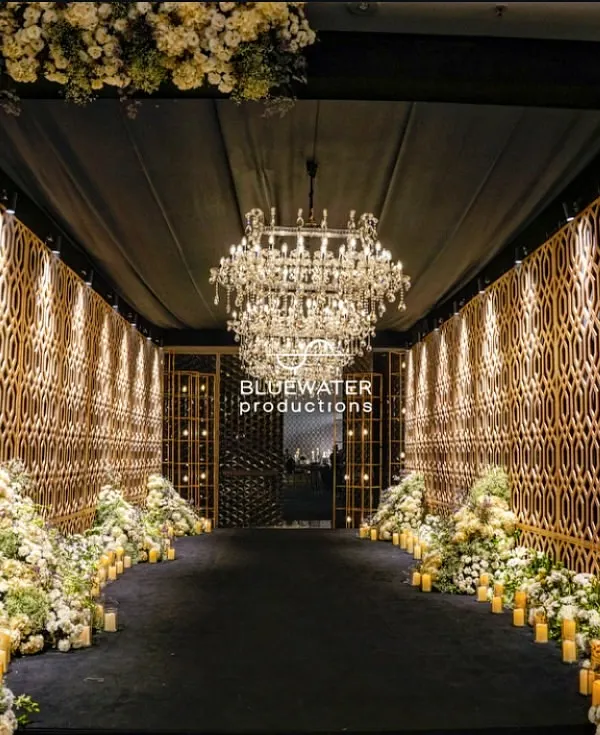 We call this a pre-party runway. A dramatic entrance tunnel with golden geometric walls, a grand chandelier, and a carpet of white florals leading guests to the Sangeet.