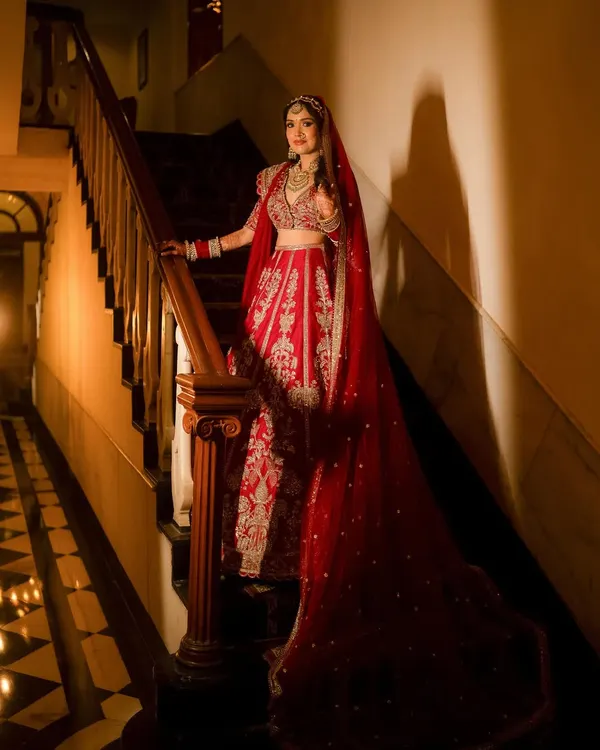 A stunning portrait of the bride on a grand wooden staircase. The dramatic lighting highlights the intricate details of her red lehenga and the elegance of her pose.