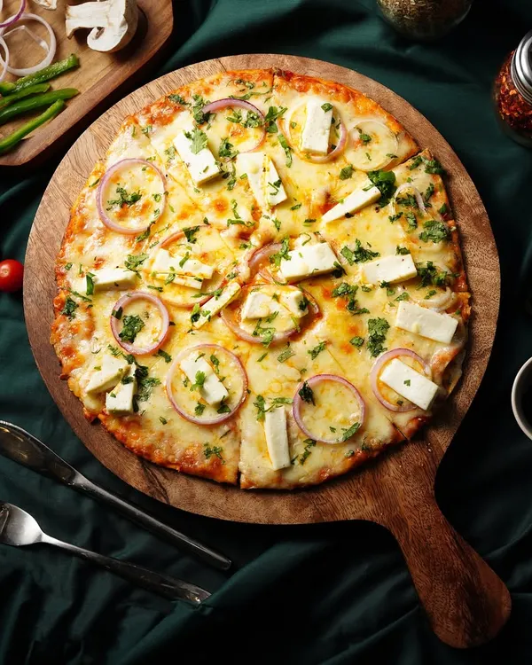 A top-down view of a delicious paneer pizza, fresh and ready to eat. We styled it with fresh ingredients on the side to emphasize its quality.