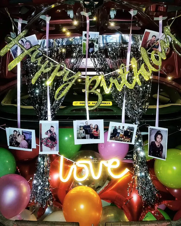A fully decorated car trunk for a birthday surprise, complete with balloons, a photo garland, fairy lights, and a 'love' neon sign.