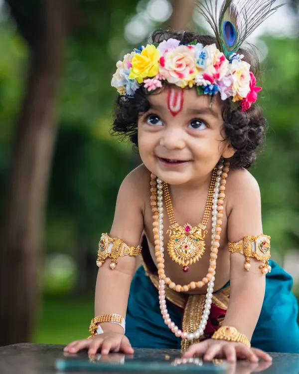 A close-up portrait of a baby boy dressed as little Krishna, complete with a floral crown, peacock feather, and traditional jewelry, smiling sweetly against a natural green background.