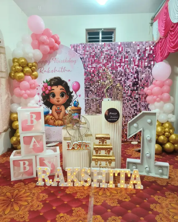 This first birthday setup for Rakshitha features a pink shimmer wall and a Moana-inspired baby character backdrop. The decor is completed with a marquee '1', 'BABY' blocks, and light-up letters for the name.