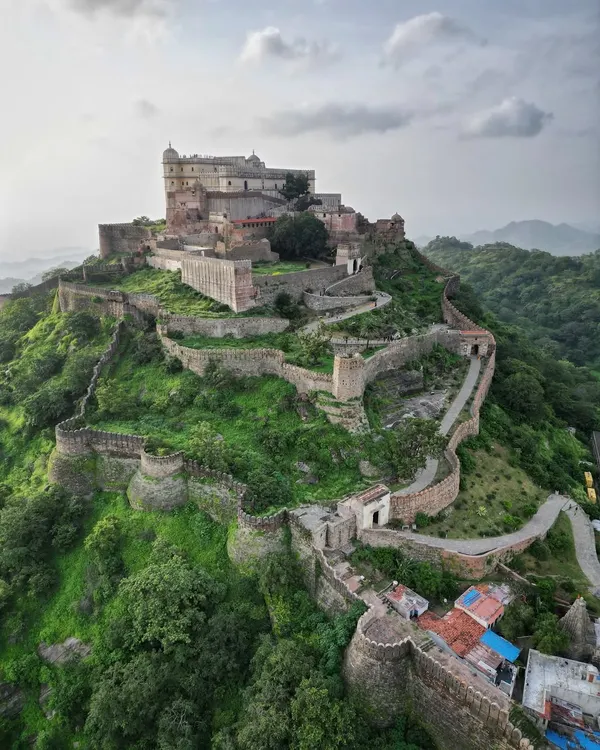 A majestic view of Kumbhalgarh Fort in Rajasthan, a UNESCO World Heritage site. The drone captures the main palace complex perched atop a hill, with its formidable fortification walls snaking through the green landscape.