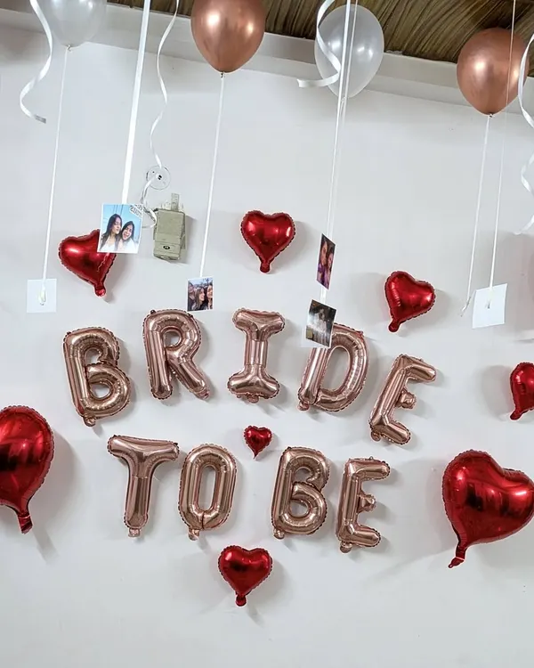 A classic 'BRIDE TO BE' room decoration featuring rose gold foil letters, red heart balloons, and photos of the bride with her friends hanging from the ceiling.
