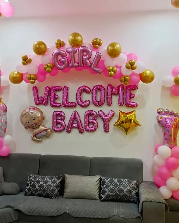 A classic "Welcome Baby" decoration for a home living room. This setup uses pink and gold balloons, foil letters, and baby-themed props to create a festive atmosphere for welcoming a baby girl.