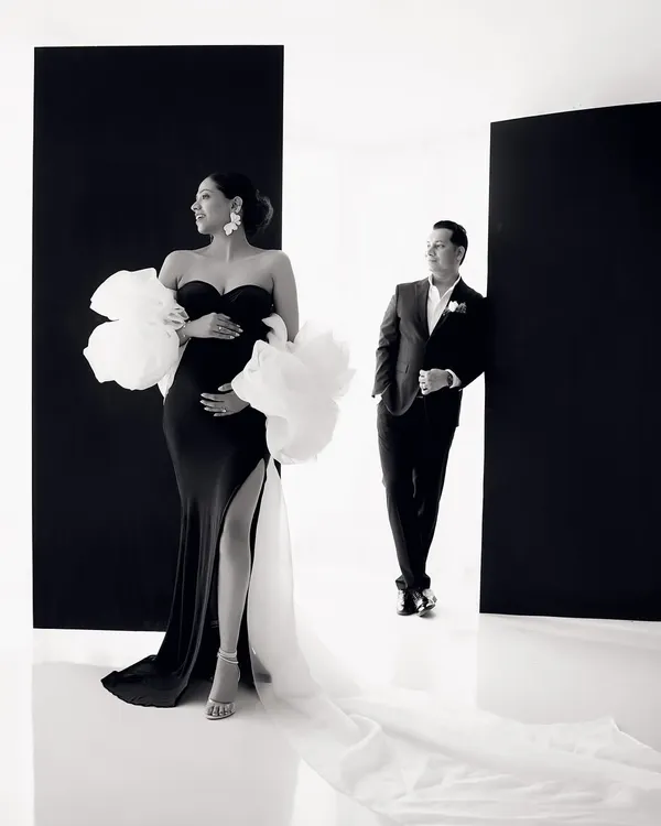 A moment of high fashion maternity. The expecting mother looks absolutely breathtaking in a black gown with dramatic white floral sleeves, creating a striking silhouette against the minimalist backdrop.