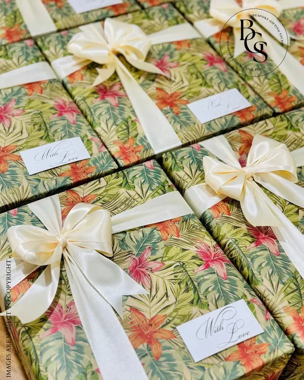 A set of gifts wrapped in a lush, tropical-print paper. The vibrant green and pink floral design is balanced with a classic cream satin bow and a simple "With Love" tag, perfect for a destination wedding or summer event.
