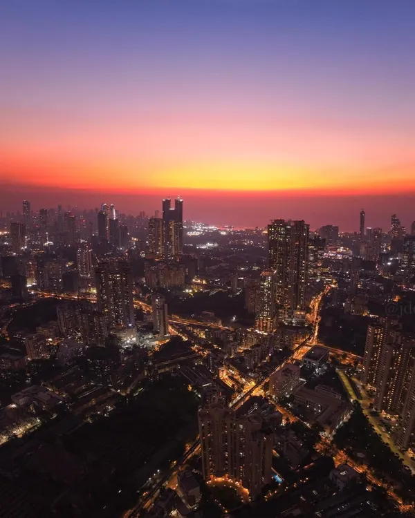 A wide-angle drone shot of the Mumbai skyline at dusk, with the city lights beginning to twinkle under a sky painted in shades of orange and purple.