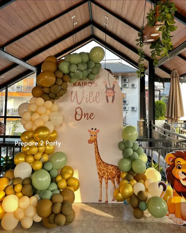 A "Wild One" jungle safari theme, perfect for a first birthday. This setup features an arched backdrop with a giraffe, a lush organic balloon garland in earthy tones, and a cute lion cutout.