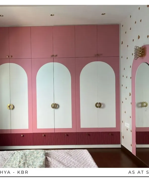 The centerpiece of this girl's room is the custom wardrobe, featuring a playful design with pink and white arched panels and unique gold hardware. The heart-patterned wallpaper adds a touch of whimsy.