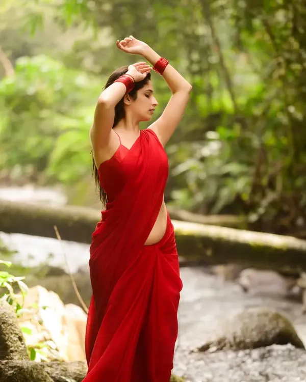 A stunning portrait in a vibrant red saree, set against the lush greenery of a forest stream. The contrast between the red and green is visually striking, creating a powerful, elemental image.