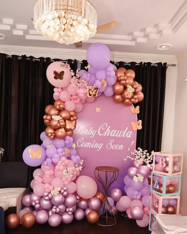 A beautiful butterfly theme for a baby shower, featuring a custom "Baby Chawla Coming Soon" backdrop. This setup uses a mix of lavender, pink, and metallic rose gold balloons, accented with delicate butterfly cutouts and floral details for an elegant look.