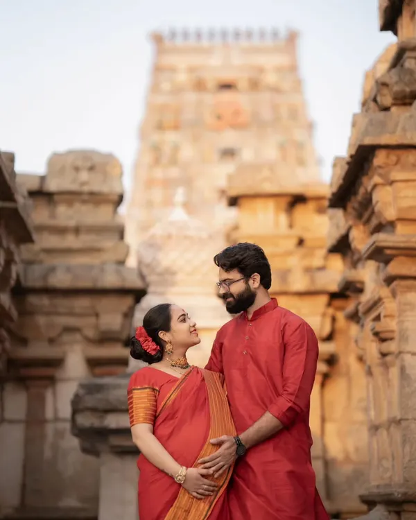 An expecting couple dressed in traditional red attire, sharing a loving gaze against the magnificent backdrop of a South Indian temple.