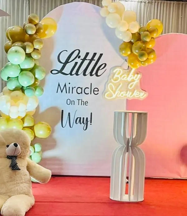 A sweet and simple baby shower setup with the theme "Little Miracle On The Way". The decor features pastel balloon clusters, a large teddy bear, and a neon sign.
