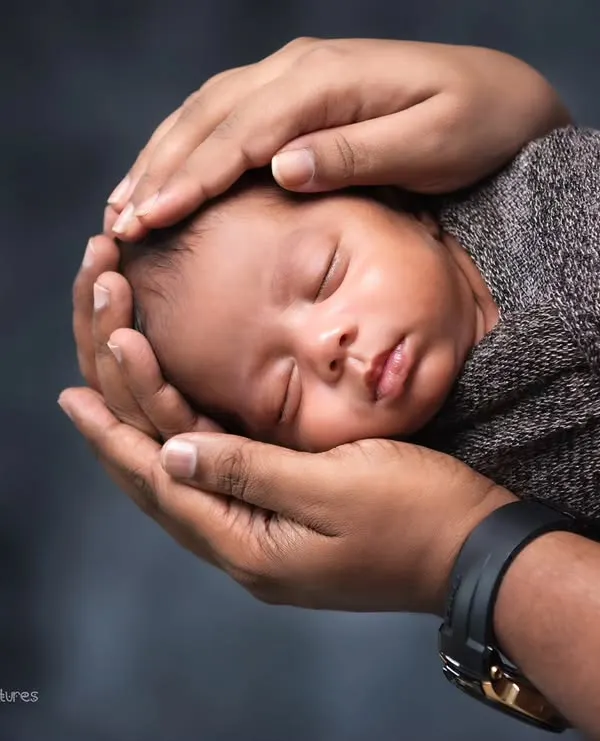 A perfect example of a safe and classic newborn pose. The baby is held securely in their parent's hands, swaddled in a neutral wrap against a dark background.