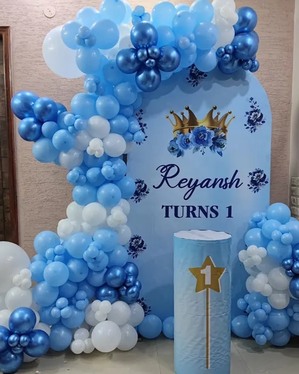 Here is a classic 'Prince' theme for a first birthday celebration. The setup includes a personalized backdrop with the baby's name, 'Reyansh', and a crown motif. The balloon arch uses various shades of blue and white, with chrome blue accents for a royal touch.