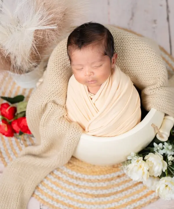 A classic and peaceful newborn portrait. This little one is perfectly swaddled and sleeping soundly in a bowl prop, surrounded by soft textures and delicate flowers for a timeless look.