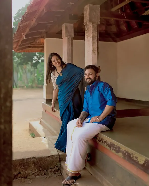 A playful, candid moment on the porch of a traditional South Indian house. The woman's blue saree and the man's mundu create a beautiful, authentic look.