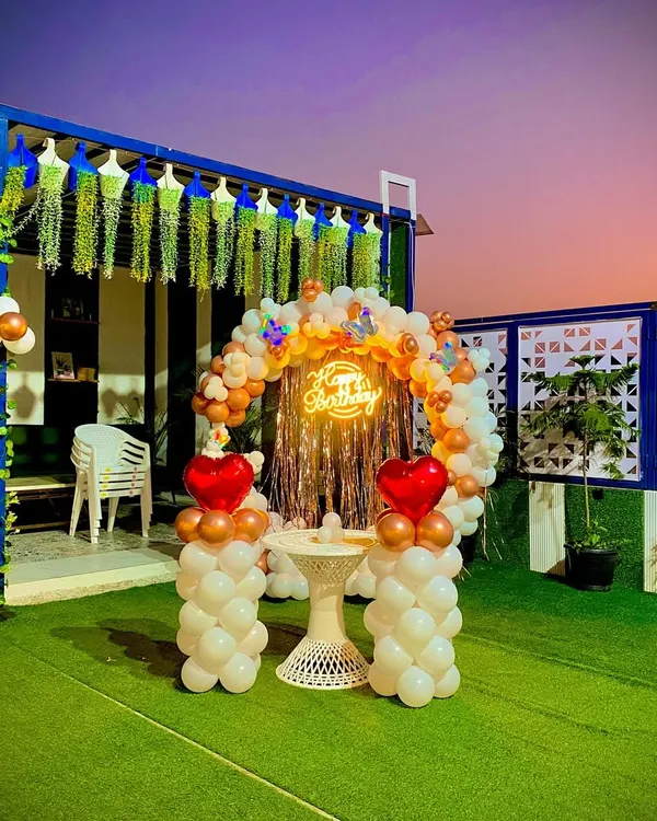 A gorgeous terrace birthday party setup at dusk. The circular balloon arch in white and bronze, accented with heart-shaped balloons, creates a stunning backdrop against the evening sky.