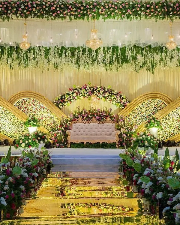 An opulent reception stage featuring golden arched panels filled with dense white flowers. The cascading floral arrangements and a grand central sofa create a look of pure elegance and luxury for a premium wedding event.