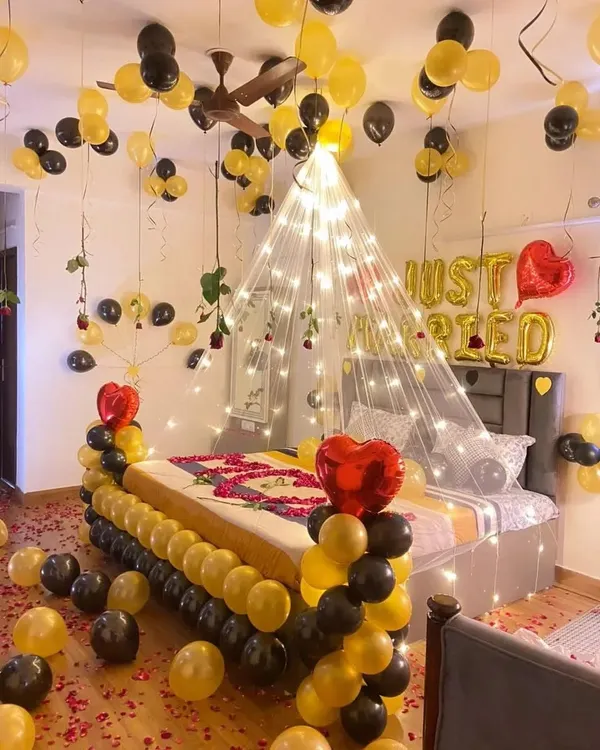 A "Just Married" first-night room decoration featuring a fairy light canopy over the bed, black and gold balloons, and scattered rose petals.