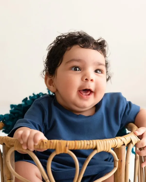 A baby with beautiful curly hair smiles brightly while sitting in a rustic wicker basket, a perfect example of a joyful sitter session.
