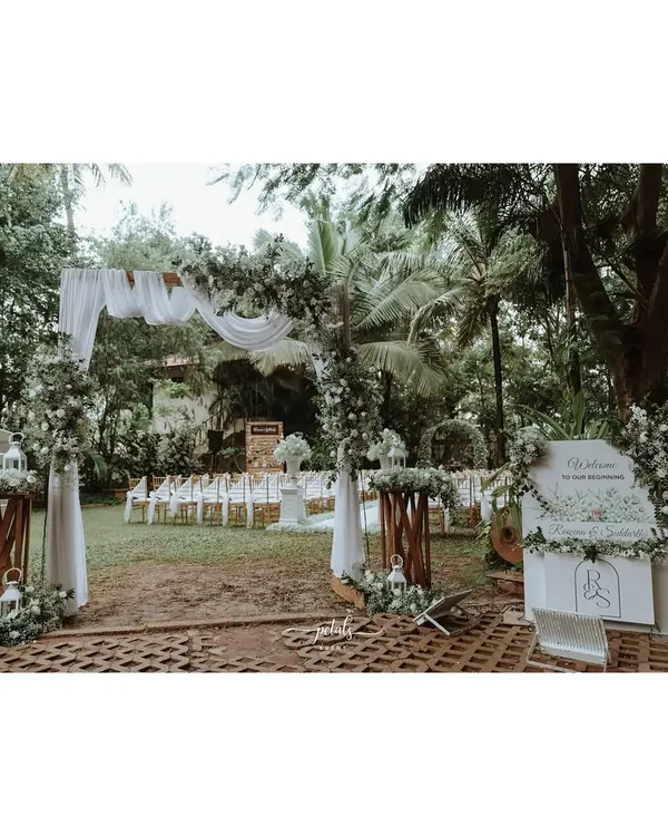 A graceful entrance for a white wedding, featuring a draped arch adorned with baby's breath and greenery. A personalized welcome sign and floral arrangements on wooden stands complete the elegant look.
