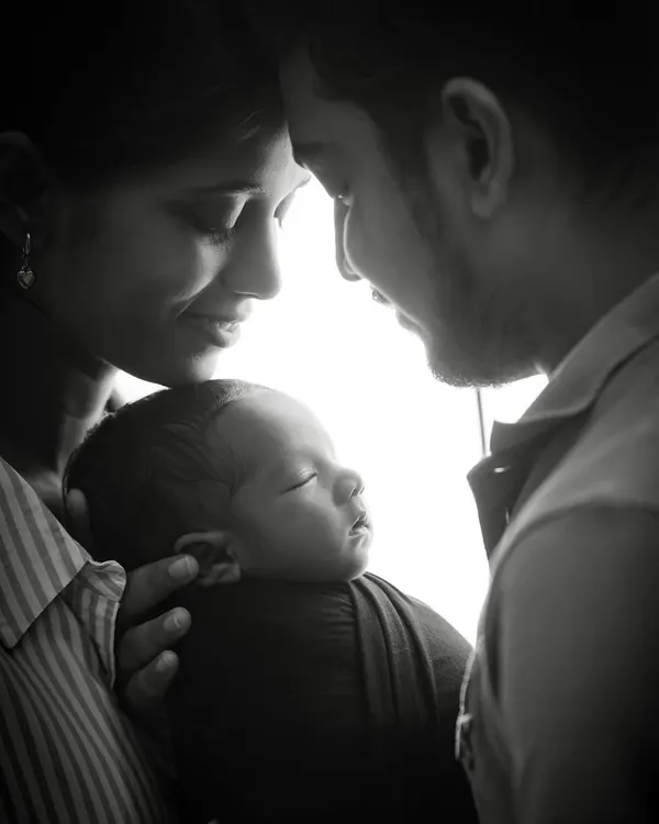 A powerful black and white portrait capturing the complete family unit. The parents gaze lovingly at their newborn, creating a silhouette effect that emphasizes their deep connection and the beginning of their new life together.