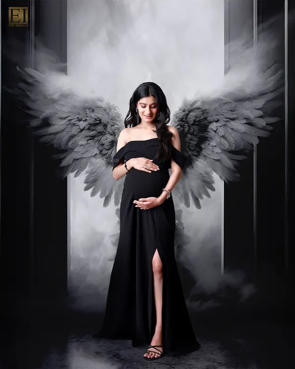 A dramatic and powerful portrait featuring a mom-to-be in a black gown with artistic angel wings, set against a moody, smoky backdrop.