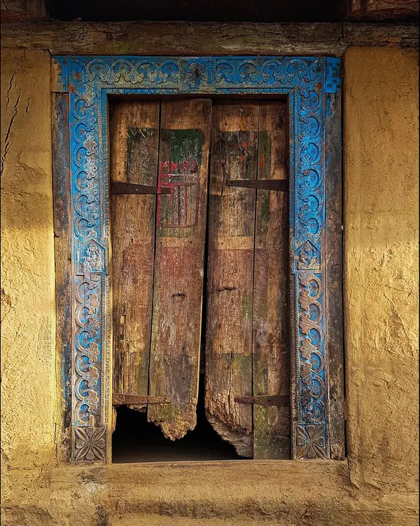 A weathered wooden door framed by an ornate, hand-carved blue border stands out against a yellow wall. The decay at the bottom hints at the countless seasons this beautiful entrance has endured.