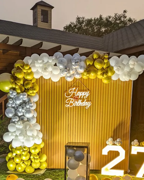 A daytime view of the 27th birthday garden party setup, showing the gold, silver, and white balloon arch against the wooden backdrop.
