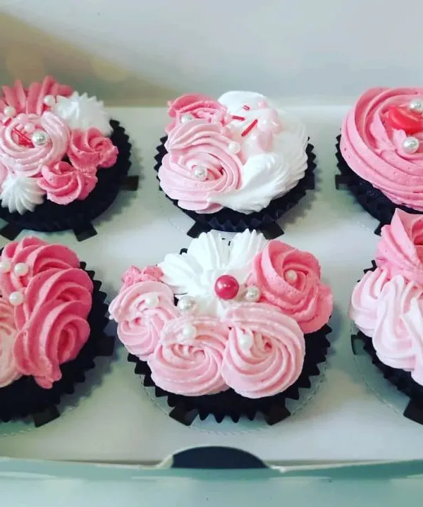 A close look at a box of six chocolate cupcakes, each topped with intricate pink and white vanilla frosting roses and delicate pearl sprinkles. Our BakeStars craft these preservative-free desserts by hand, making them perfect for birthdays or just a little self-love.
