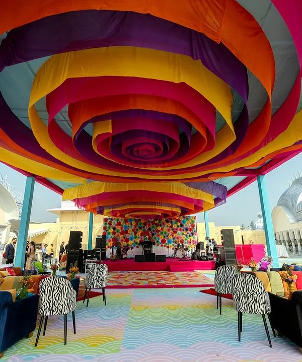 A whirlwind of color at a Carnival-themed pre-wedding event. The ceiling is a spiral of vibrant drapes, leading to a stage with a backdrop of colorful paper fans, creating an atmosphere of pure joy and celebration.