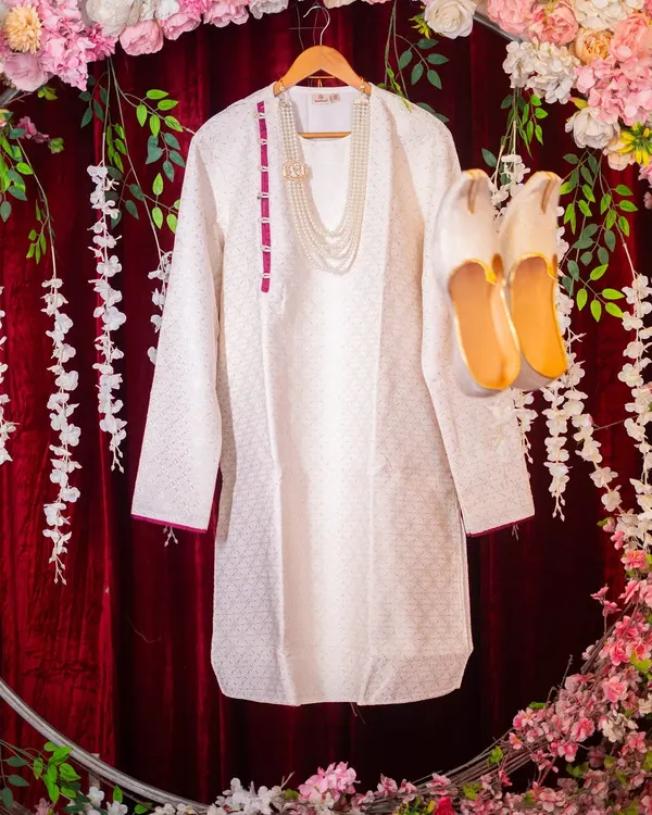 An artistic flat-lay of the groom's wedding attire, including his white kurta, pearl necklace, and traditional shoes (mojari), beautifully arranged.