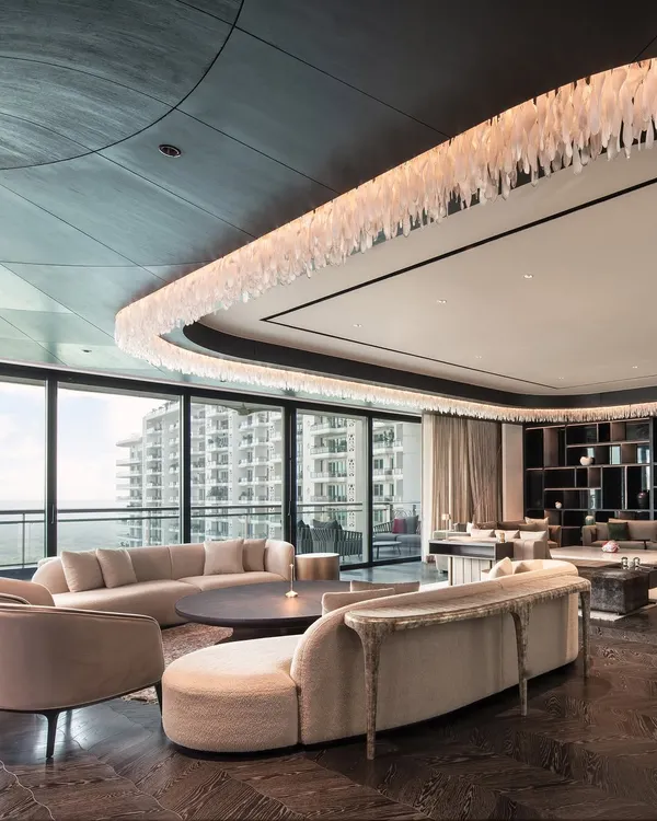 An expansive living area in a DLF Camellias apartment, where curved sofas soften the grand scale of the room. The custom furniture pieces are arranged to create multiple zones for conversation and relaxation, all unified by a neutral palette and luxurious textures.