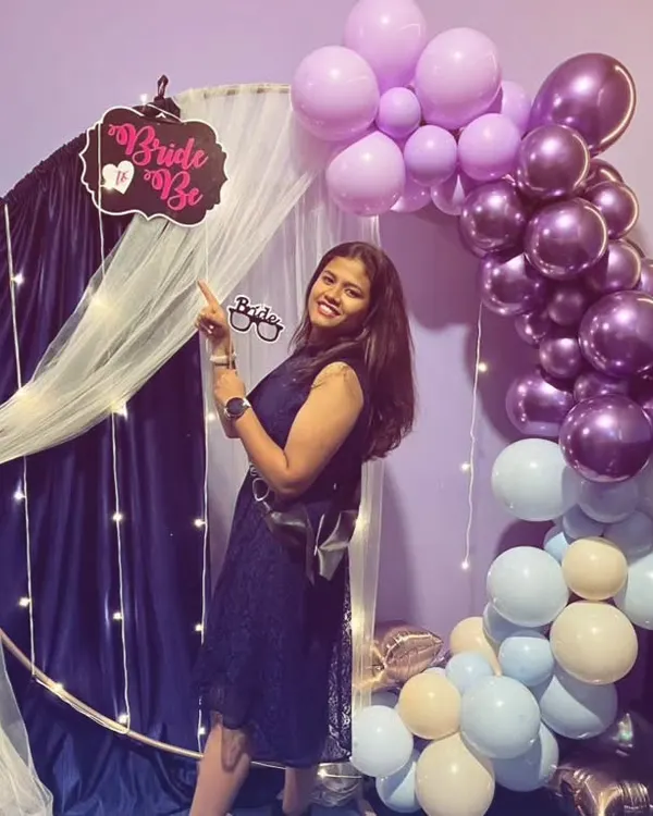 The bride-to-be posing with a 'Bride to Be' sign in front of her custom party decorations. The purple and blue balloon theme was chosen by her friends to surprise her.