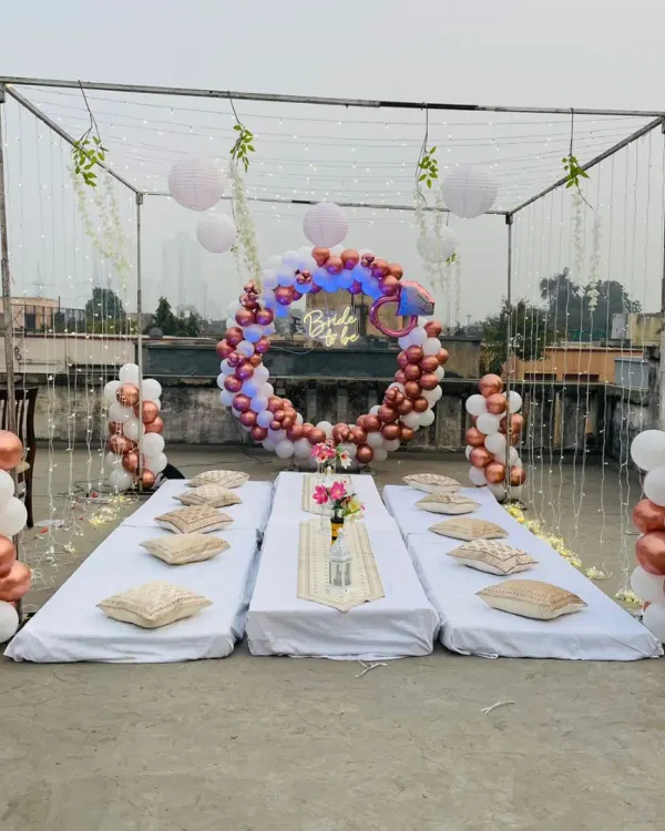 A beautiful 'Bride to be' terrace party setup. This features a metal frame with fairy lights, a circular balloon arch in rose gold and white, and a cozy low-seating arrangement.